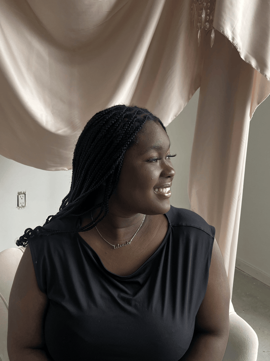 A dark skin woman sits in the white chair with a pink backdrop wearing a silver necklace that reads, beloved, with a heart.